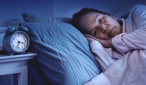 A woman lies awake in bed next to an alarm clock reading half past three