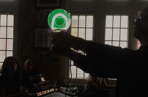A man hold a green glowing ball with a white ring inside it, silhouetted against a window.