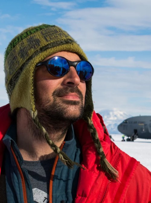 A man in sunglasses and a wooly hat with a snowy landing strip behind