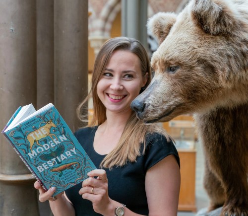 A woman reading a book while a bear looks over her shoulder. The book is The Modern Bestiary.